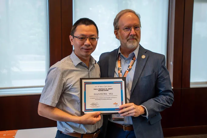 Two individuals holding a certificate between them for a photo. One person is wearing a plaid shirt and glasses; the other is in a light blue shirt and has a beard. They are both smiling and standing for a photo. The certificate mentions a recipient named Qiusheng Wu.