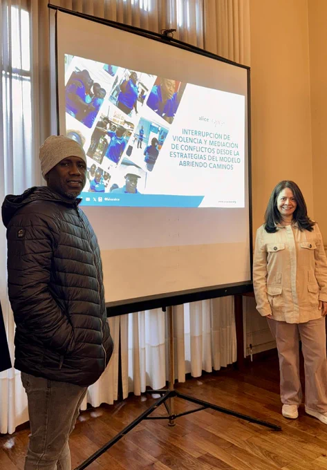 Two session facilitators standing on either side of a projector screen. The person on the left is wearing a black puffer jacket and a white beanie. The person on the right is wearing a button down jacket and beige slacks.