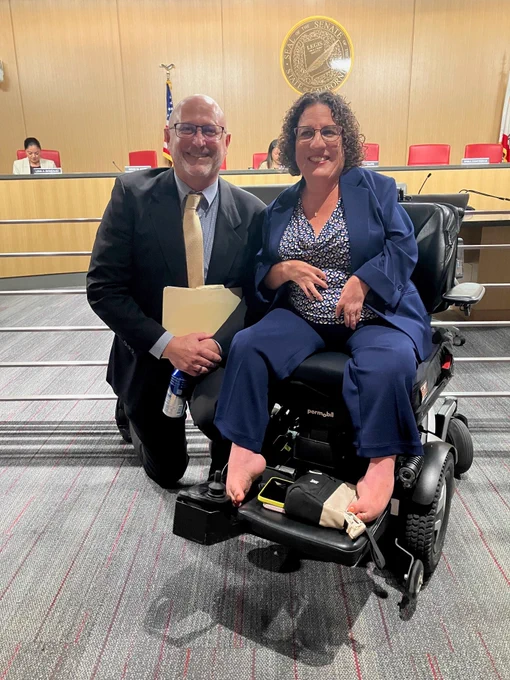 Directors Pete Cervinka kneeling next to Director Rutledge seated in a motorized wheelchair, both smiling at the camera at their Senate Rules Committee confirmation hearing, with a United States flag and government seating in the background. 