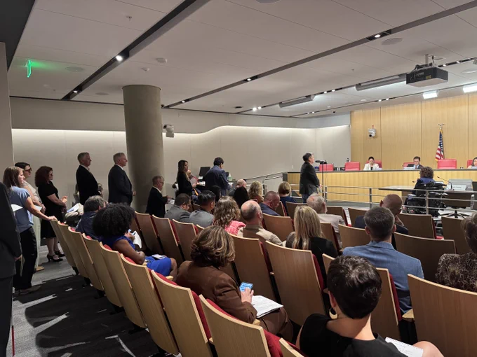 Audience members seated around a group of individuals standing in line to speak at a microphone, attending a hearing inside a modern courtroom setting, with an American flag in the background.