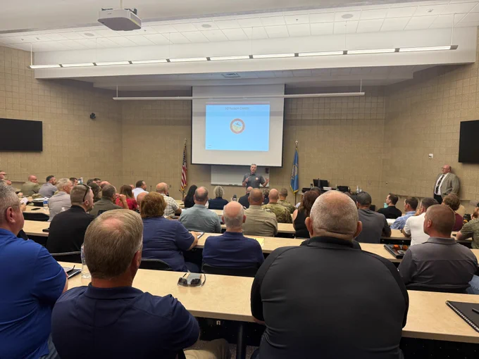 A full classroom of emergency management personnel facing a speaker at the front of the room, with a presentation slide titled "SD Fusion Center" projected on the screen. American and South Dakota flags are visible near the speaker.