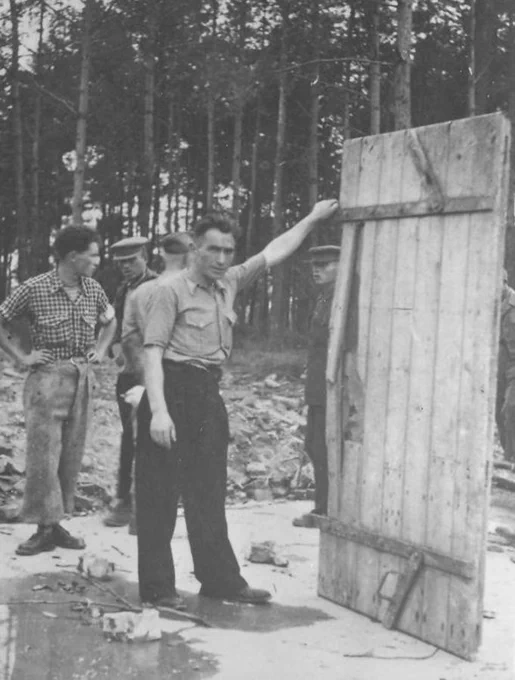 Three individuals in a wooded area examining a large, wooden gas-tighed door - remains of gas chamber and crematorium V at the former Auschwitz II-Birkenau site.
