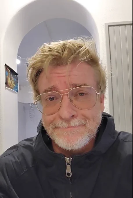 Picture of Rhys Darby, blonde hair, dad glasses, black zip up jacket smiling at the camera