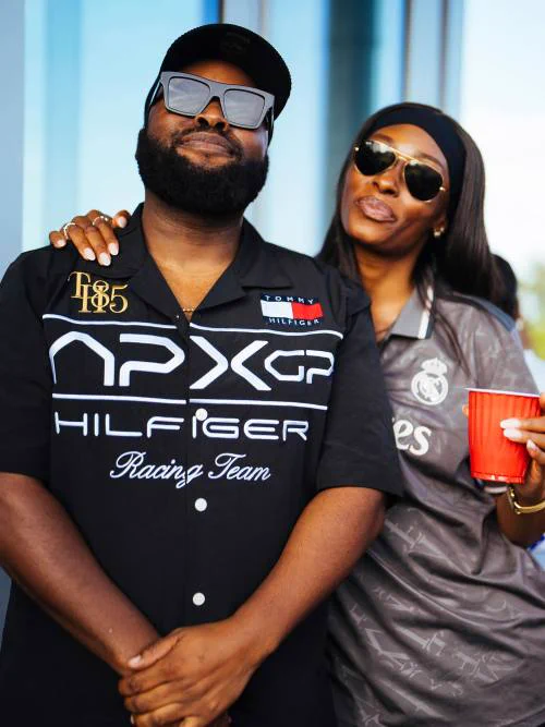 Picture of Samson standing smiling at the camera with hands in front of him wearing a black shirt that says APXXGP Hilfiger Racing Team. On his right, a female presenting person has her hand on his shoulder (apologies I don't know her name) she is also smiling at the camera holding a red cup.