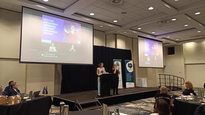 Elise Mather stands at a lectern presenting at the AFDO Disability Advocacy Conference. An Auslan interpreter is beside them. A large screen above shows the speaker's slides and a live video feed with captions. The AFDO and Business Inclusion and Diversity Service banners are visible on stage.