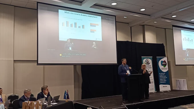 Peter Bacon stands at a lectern presenting at the AFDO Disability Advocacy Conference. An Auslan interpreter is beside them. A large screen above shows the speaker's slides and a live video feed with captions. The AFDO and Business Inclusion and Diversity Service banners are visible on stage.