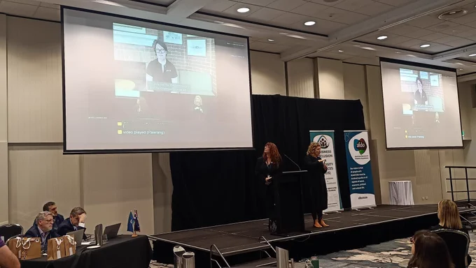 Angie Elphick stands at a lectern presenting at the AFDO Disability Advocacy Conference. An Auslan interpreter is beside them. A large screen above shows the speaker's slides and a live video feed with captions. The AFDO and Business Inclusion and Diversity Service banners are visible on stage.