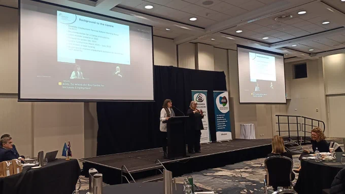 Lucy Macali stands at a lectern presenting at the AFDO Disability Advocacy Conference. An Auslan interpreter is beside them. A large screen above shows the speaker's slides and a live video feed with captions. The AFDO and Business Inclusion and Diversity Service banners are visible on stage.