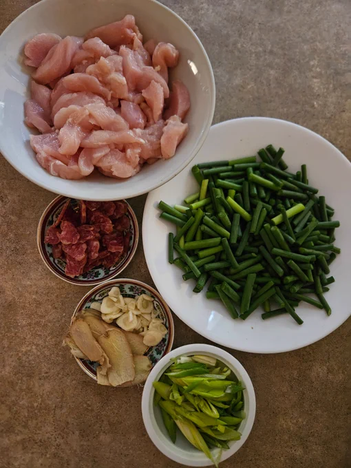 Ingredients from top of a bowl of chicken breast slices, a white soup plate with garlic scape pieces, a small saucer with sliced Chinese sausage, a small dish with ginger slices & chopped garlic, a small bowl with spring onion slices.