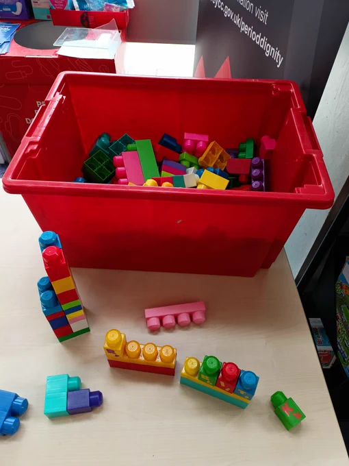 Photograph of a red tub filled with Duplo Lego blocks on a wooden table. Other blocks are arranged on the table in front of the tub.