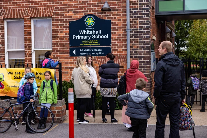 Children and parents, some on foot and some on cycles, gather at the school gates of a primary school. 