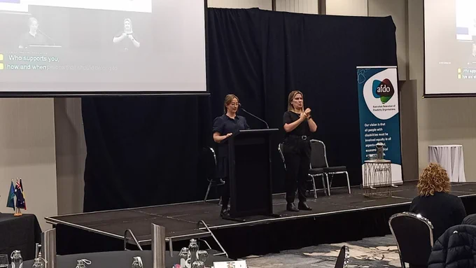 Brittany Pantano from Enliven Community speaks from a lectern during her session at the AFDO Disability Advocacy Conference. An Auslan interpreter stands beside her. A large screen behind them shows a captioned presentation, and the AFDO banner is visible on stage.