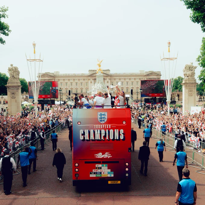 Wide photo of Englnd open top bus with players holding the EURO 2025 trophy aloft in front of Buckingham Palace. 