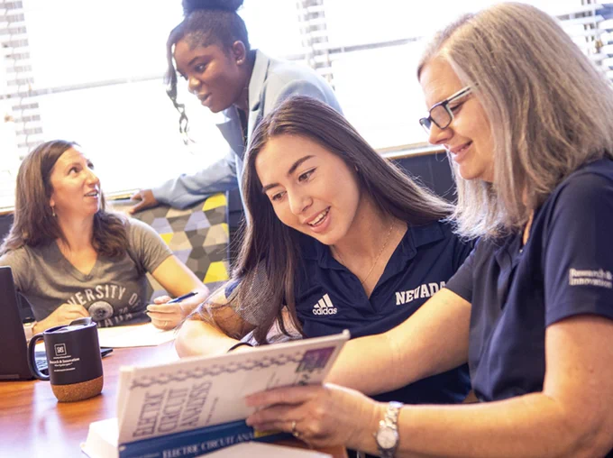 Four individuals are engaged in a discussion around a table, with one of them showing a document to another. They are in a room with a view of a cityscape outside. Two are wearing University of Nevada branded apparel.