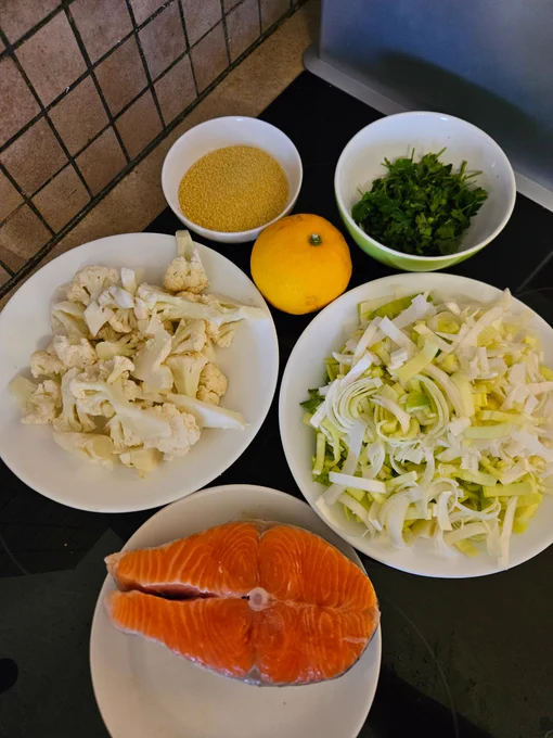 Ingredients clockwise from left of a plate with cauliflower pieces, a small bowl of dry couscous, a lemon, a small bowl of chopped parsley, a bowl of sliced leek, a small white plate with a salmon steak.