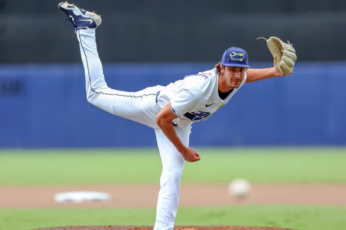 Paul Gervase follows through after a pitch with his right leg high in the air. He wears an all-white uniform with a light brown glove on his left hand.