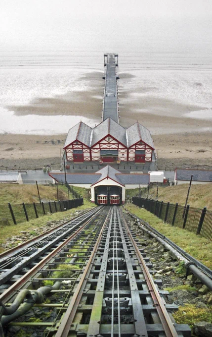 A photograph of steep funicular railway tracks descending towards a red and white pavilion, leading to a long pier extending over a sandy beach.