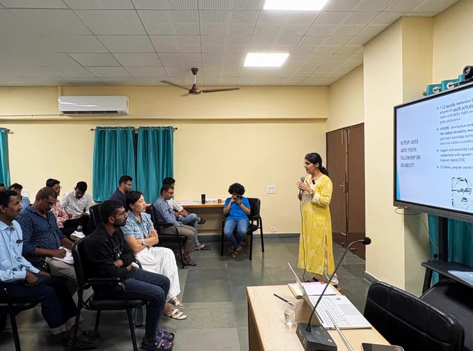 NCPEDP's Vamika Gupta is standing with a mic on her hand, addressing the people present in front of her. There is a white screen behind her.