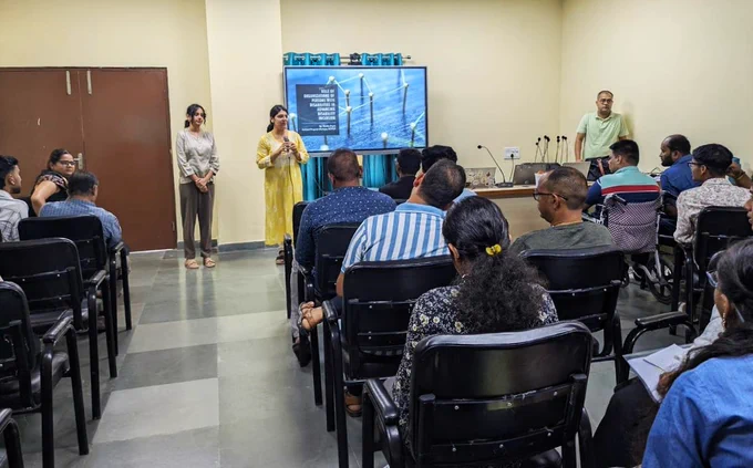 NCPEDP's Vamika Gupta is standing with a mic on her hand, addressing the people present in front of her. There is a white screen behind her. Ananya Sharma is next to her