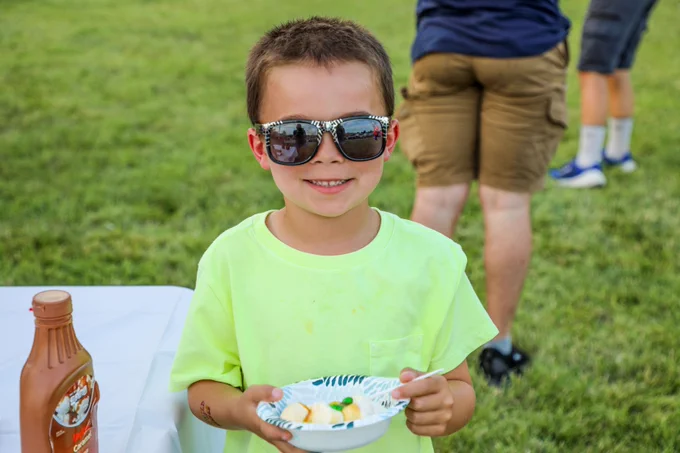 A young boy wearing zebra-striped sunglasses and a neon yellow shirt smiles while holding a bowl of ice cream at an outdoor event. A bottle of caramel sauce sits on the nearby table.