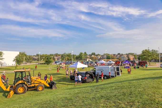 A wide view of a community event at a park showing families walking around tents, city vehicles including a backhoe, fire truck, and CSI van. The event is set on green grass under a blue sky with suburban homes and cars in the background.
