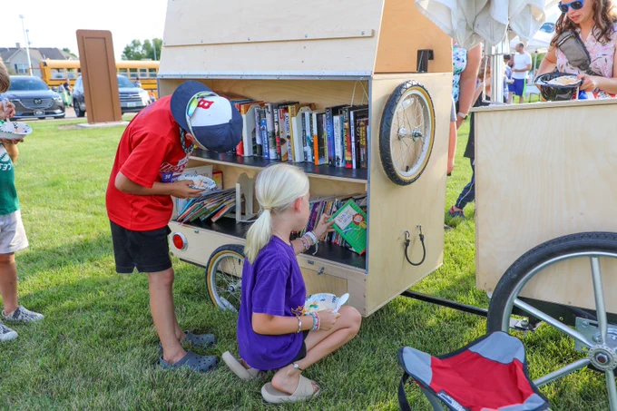 Two children look through books on a mobile wooden library cart with shelves of books for kids and adults. The cart has bicycle wheels and is set up on a grassy field during a sunny outdoor event.