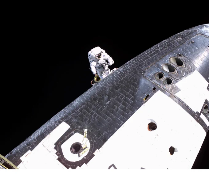 NASA Astronaut Stephen Robinson, seen in his protective spacesuit, is poised along Discovery's dark underbelly during STS-114's third spacewalk. He carefully works to remove protruding gap fillers from between the orbiter's heat-shielding tiles. Behind him is the blackness of space. Date: August 3, 2005. Credit: NASA