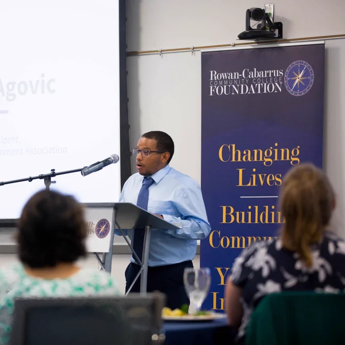 A student speaks at a podium during an event at Rowan-Cabarrus Community College. Audience members are seated at tables in front of him.