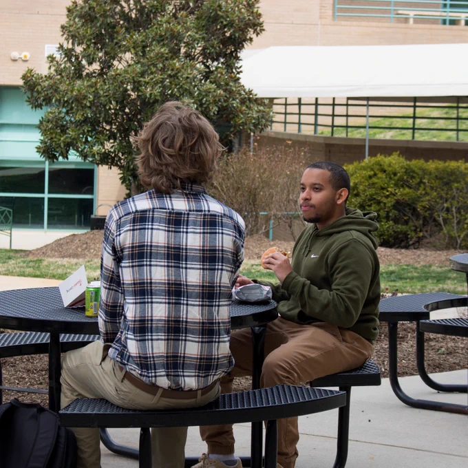 Two students sit at a black metal picnic table outside on a college campus. One wears a green hoodie and holds a sandwich, while the other wears a plaid shirt with their back to the camera. A modern campus building and trees are in the background.