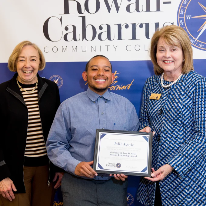 A student stands smiling between two college officials at a Rowan-Cabarrus Community College event. He holds a framed award certificate recognizing him as the recipient of the Governor Robert W. Scott Student Leadership Award. A blue and white Rowan-Cabarrus backdrop is behind them.