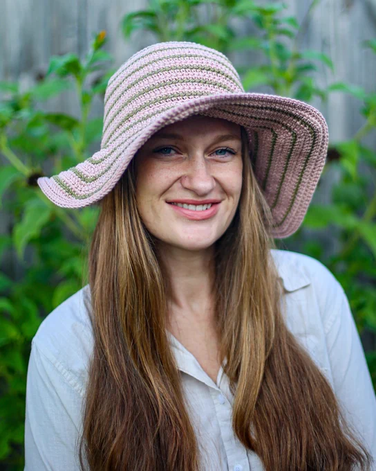 Person wearing a wide-brimmed pink and green striped crochet hat with long brown hair and a white button-up shirt, standing outdoors.