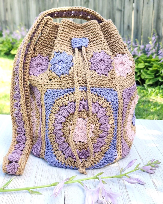 Handcrafted crochet drawstring bag featuring pastel purple, pink, and beige floral motifs on a white wooden surface outdoors.