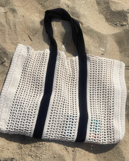 Beige crochet tote bag with black handles resting on sandy beach with faint shadows.