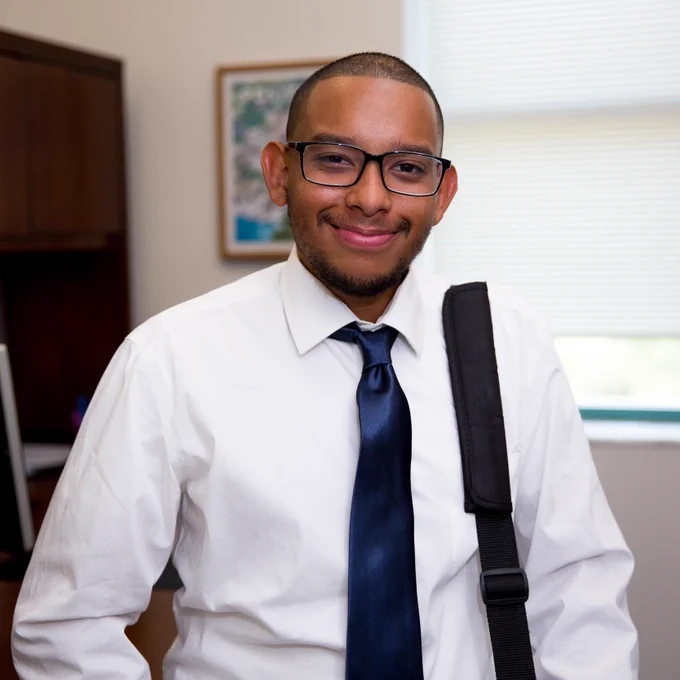A young man wearing a white shirt, navy blue tie, and glasses stands smiling in an office setting with a computer and framed artwork in the background. A shoulder strap is visible, suggesting he is carrying a bag.