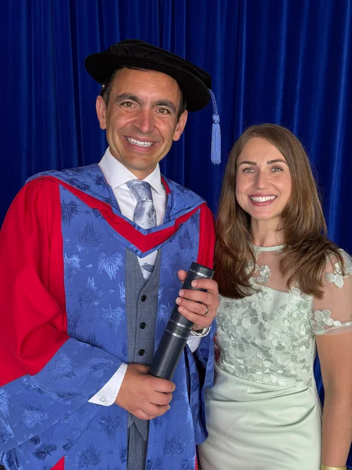 A photograph of Steven Mifsud MBE posing for a photo alongside his wife, Direct Access Co-Director Jude Mifsud. Steven is wearing a blue and red graduation robe and holding a black tubular scroll containing his doctorate. Jude is wearing a half-sequined silver dress. Both are smiling.