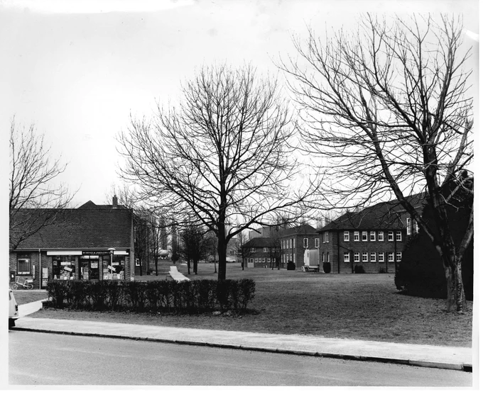 Black and white image. The left hand side of the shot shows the end of the row of shops on Central Avenue, Wharley End. To the right there are two trees and a grassy lawn stretching backwards, with a view of the Stafford Cripps building in the distance.