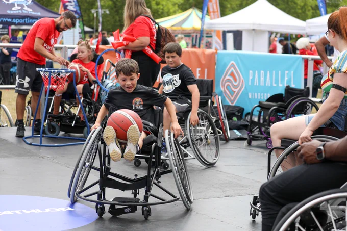 A photo in the Parallel Actice lifestyle zone which has multiple people on sport wheelchairs and a basketball ring on the left. the focus of the image is on a little boy sat in a sports chair with a black 'titans basketball' t shirt on and a basketball on his knees. His tongue is stuck out the side as he concentrates with a big grin! In the background are other tents and activities. 
