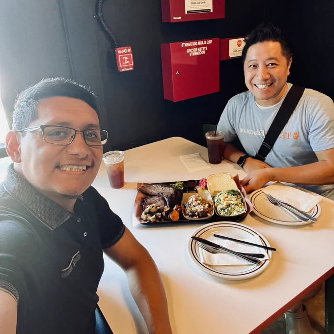 Two people are sitting at a small restaurant table with a platter of assorted barbecue dishes, including smoked meats, sides, and bread, placed between them. Each person has a drink with ice and a plate with utensils. The setting has a casual, modern vibe with dark walls, visible fire alarm boxes, and natural daylight coming in from the left. One person is wearing a black collared shirt, and the other is wearing a light gray T-shirt with a black shoulder strap.