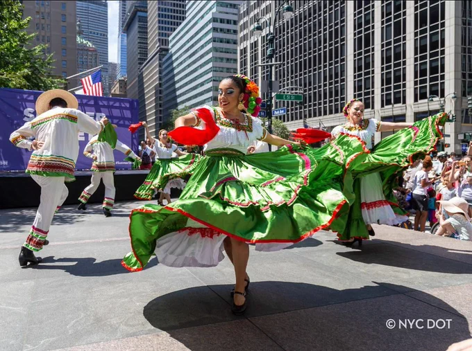 A photo of dancers in traditional dress, including green dresses swirling as they dance, performing on a NYC street. 