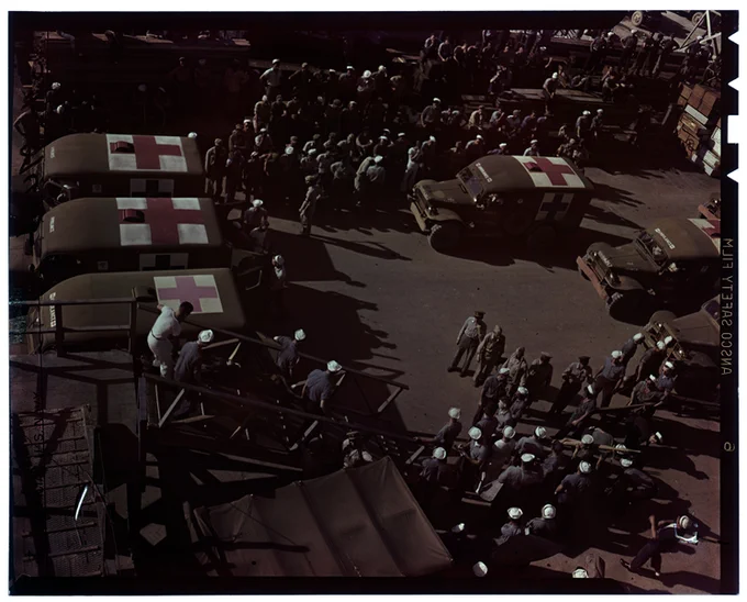 Original Caption: "Survivors of the USS Indianapolis (CA-35) being brought ashore from USS Tranquility (AH-14) at Guam. They are being placed in ambulances for immediate transfer to local hospitals." Photographer: PhoM1/c J.G. Mull. [Note: Image has been color corrected]

Several military ambulances with large red crosses on their roofs are surrounded by hundreds of sailors.  The photograph was taken from above.