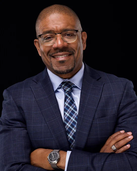 Lemichael Simmons in a blue plaid suit with glasses, standing with crossed arms.