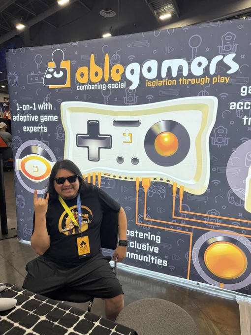 Rattlehead sitting in front of an "AbleGamers" booth display at Evo. The background banner features a large game controller graphic and text that reads: "AbleGamers: combating social isolation through play," along with phrases like "1-on-1 with adaptive game experts" and "fostering inclusive communities." Rattle is smiling, wearing sunglasses, a black t-shirt with a Mortal Kombat logo, black shorts, and a yellow event lanyard with a badge that says "EVO Exhibitor." He is making a "rock on" hand gesture.