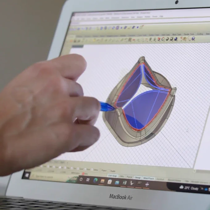 A person's hand holds a pen, pointing at a 3D heart valve model on a MacBook Air screen, showing detailed design elements.