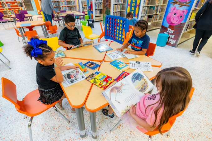 An aerial shot of a table with four children, three girls and one boy, reading various books at the library. Other books are scattered across the table as they plan to read those books next. 