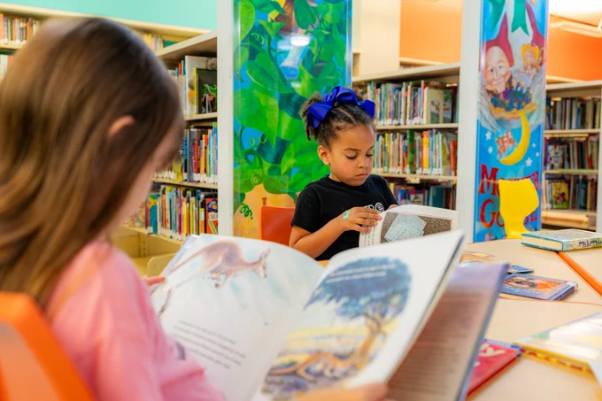 A young girl with ribbons in her hair flips through the pages of a book. She is in the library, seated at a table. In the foreground is another girl with her back turned toward us who is reading her book, which is wide open where we can see illustrations of a kangaroo and her joey.  