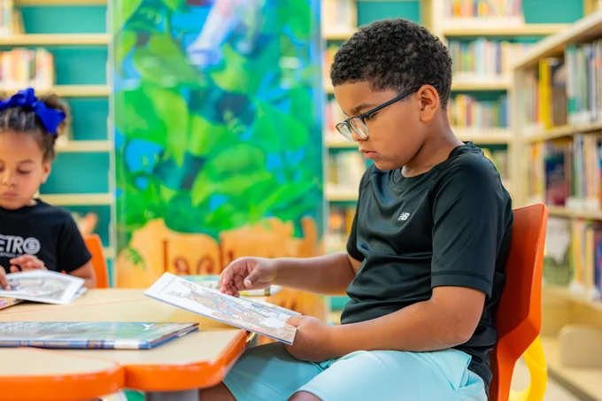 A young boy wearing glasses is reading a picture book. He is seated at a children's table in the library. In the background, a young girl with ribbons in her hair can be seen reading a book. Also in the background is a colorful wall panel and shelves filled with many books.