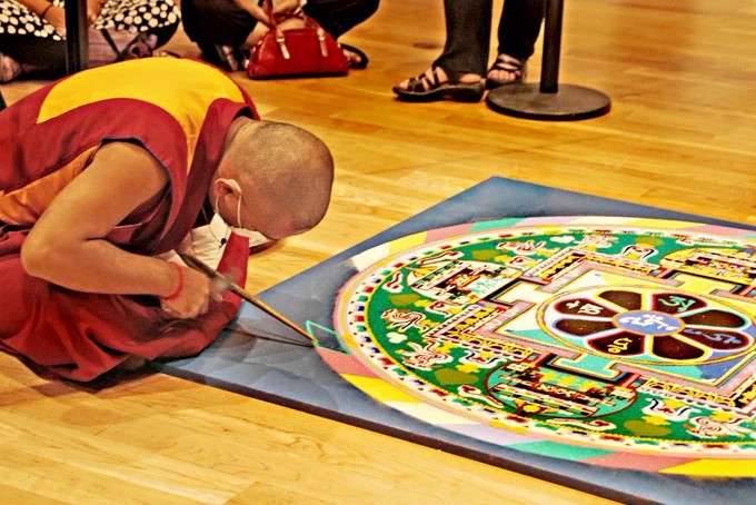 Buddhist monk in saffron robes kneeling on a wooden floor meticulously adding sand to a mandala