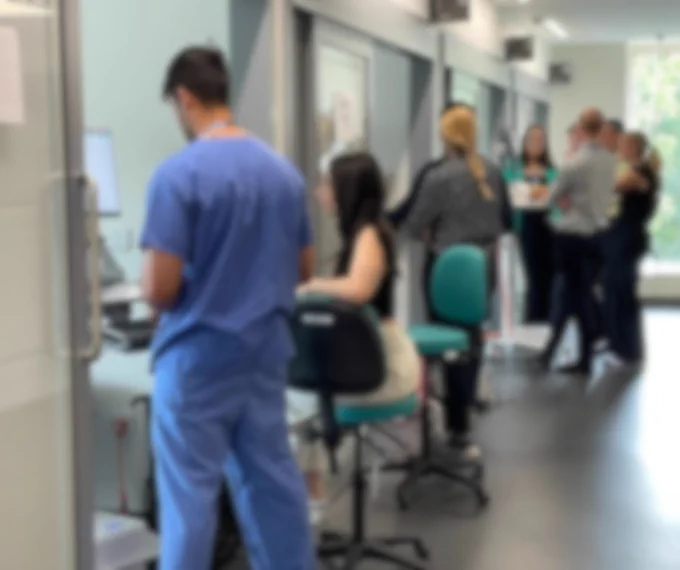 A blurred image showing several individuals in a clinical setting, with one person in medical scrubs working at a computer station and others engaging in a discussion in the background.