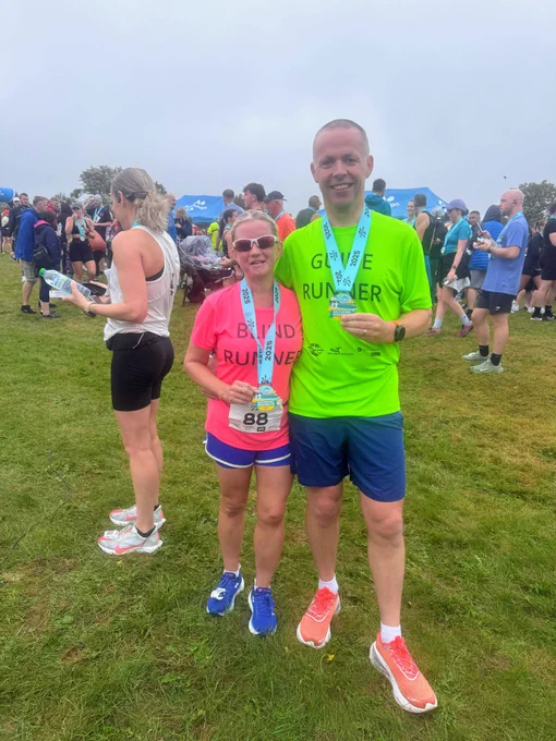 Two people standing on grass at a race event, both wearing medals around their necks. The woman on the left is wearing a bright pink shirt with the words "BLIND RUNNER" printed on it, blue shorts, and blue running shoes. She is also wearing sunglasses and holding her medal. The man on the right is wearing a neon green shirt with "GUIDE RUNNER" printed on it, blue shorts, and orange running shoes. He is smiling and holding his medal. There are other runners and participants in the background, some wearing medals and athletic gear, and a few blue tents can be seen. The atmosphere looks cheerful and celebratory, likely at the finish line of a race.