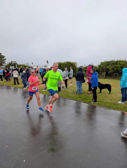 Outdoor running event on a cloudy day. In the foreground, a man and a woman are running together. The man is wearing a bright neon green shirt with the words "GUIDE RUNNER" printed on it, and the woman beside him is wearing a pink shirt with "BLIND" written on it, along with blue shorts and sunglasses. They appear to be participating as a guide and a visually impaired runner. Both are focused and mid-stride. In the background, there are several people standing on the grass, some wearing jackets and hoods, likely due to the cool, damp weather. The ground is wet, suggesting recent rain. A black dog is also visible among the spectators. The scene is lively, with a supportive atmosphere.
